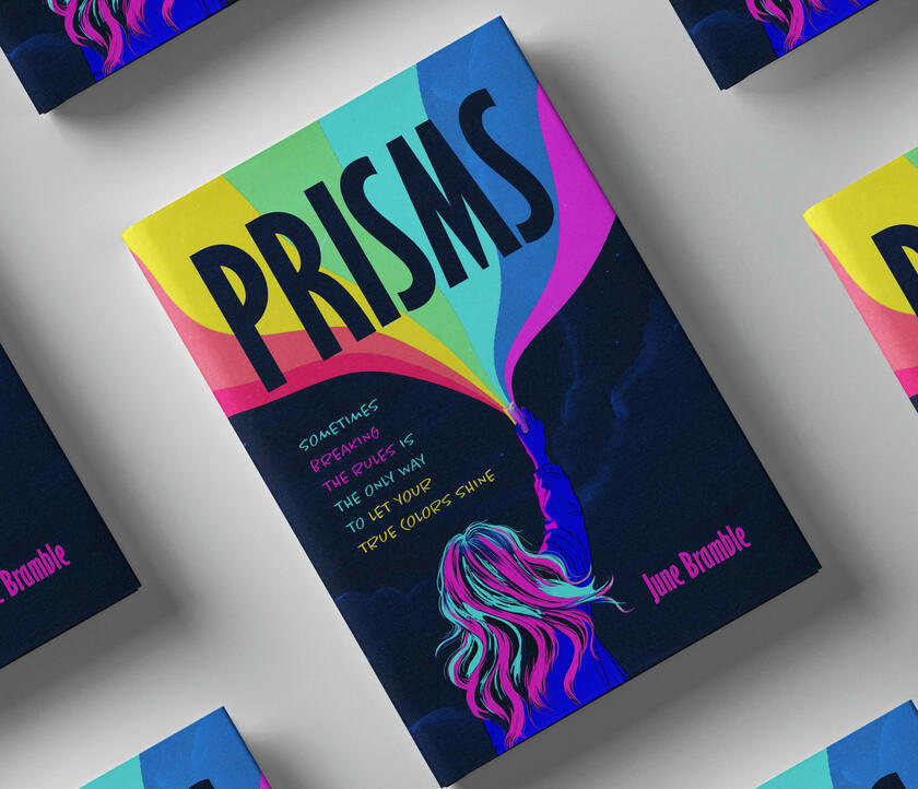 A physical copy of the book Prisms by June Bramble. On the cover, a kid with pink and blue hair holds a prism high above her, and a large rainbow shines from it.